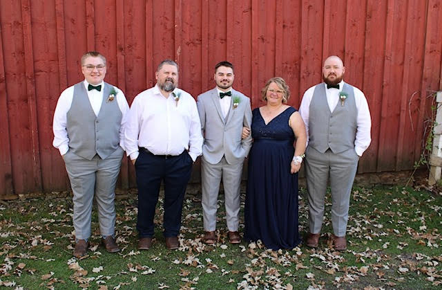 Five people pose in front of a red wooden wall, dressed in formal attire, standing on grass with scattered dry leaves.